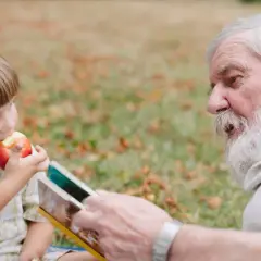 Dziadek czyta wnuczkowi książkę, a chłopiec zajada jabłko. Widać tu piękne, rodzinne powinowactwo.