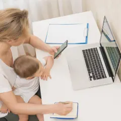 Kobieta z dzieckiem na kolanach sprawdza w telefonie, ile płatny urlop macierzyński. Obok laptop i notatnik.