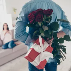 Mężczyzna z bukietem czerwonych róż i prezentem za plecami, przygotowuje niespodziankę z okazji imienin.