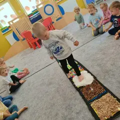 Dzieci aktywnie w żłobku ćwiczą zmysły na ścieżce sensorycznej. Maluchy z zaciekawieniem obserwują kolegę stąpającego po różnych fakturach.