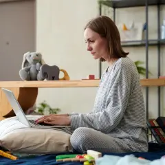 Kobieta w piżamie siedzi na łóżku, pisząc na laptopie. Obok pluszowy królik i zabawki. Przygotowuje wniosek o urlop rodzicielski.