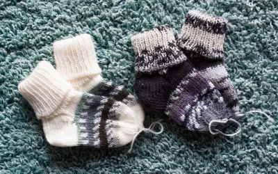 Dwa ręcznie robione, wełniane skarpetki, białe i wielokolorowe, leżą na turkusowym dywanie. Dziś, 21 października, idealne na chłodne dni.