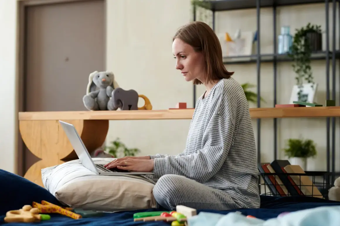 Kobieta w piżamie siedzi na łóżku, pisząc na laptopie. Obok pluszowy królik i zabawki. Przygotowuje wniosek o urlop rodzicielski.