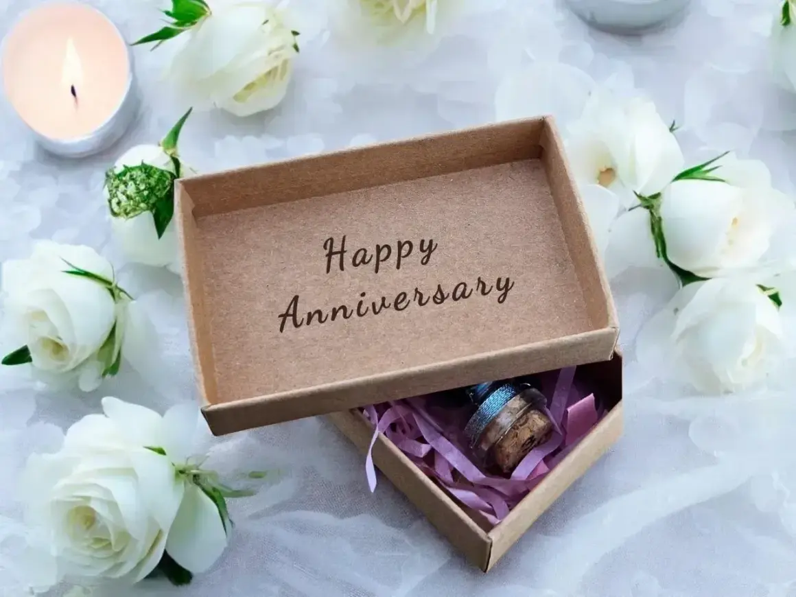 Otwarta szkatułka z napisem "Happy Anniversary" i małym prezentem w środku, otoczona białymi różami i świecą. Idealna na porcelanową rocznicę ślubu.