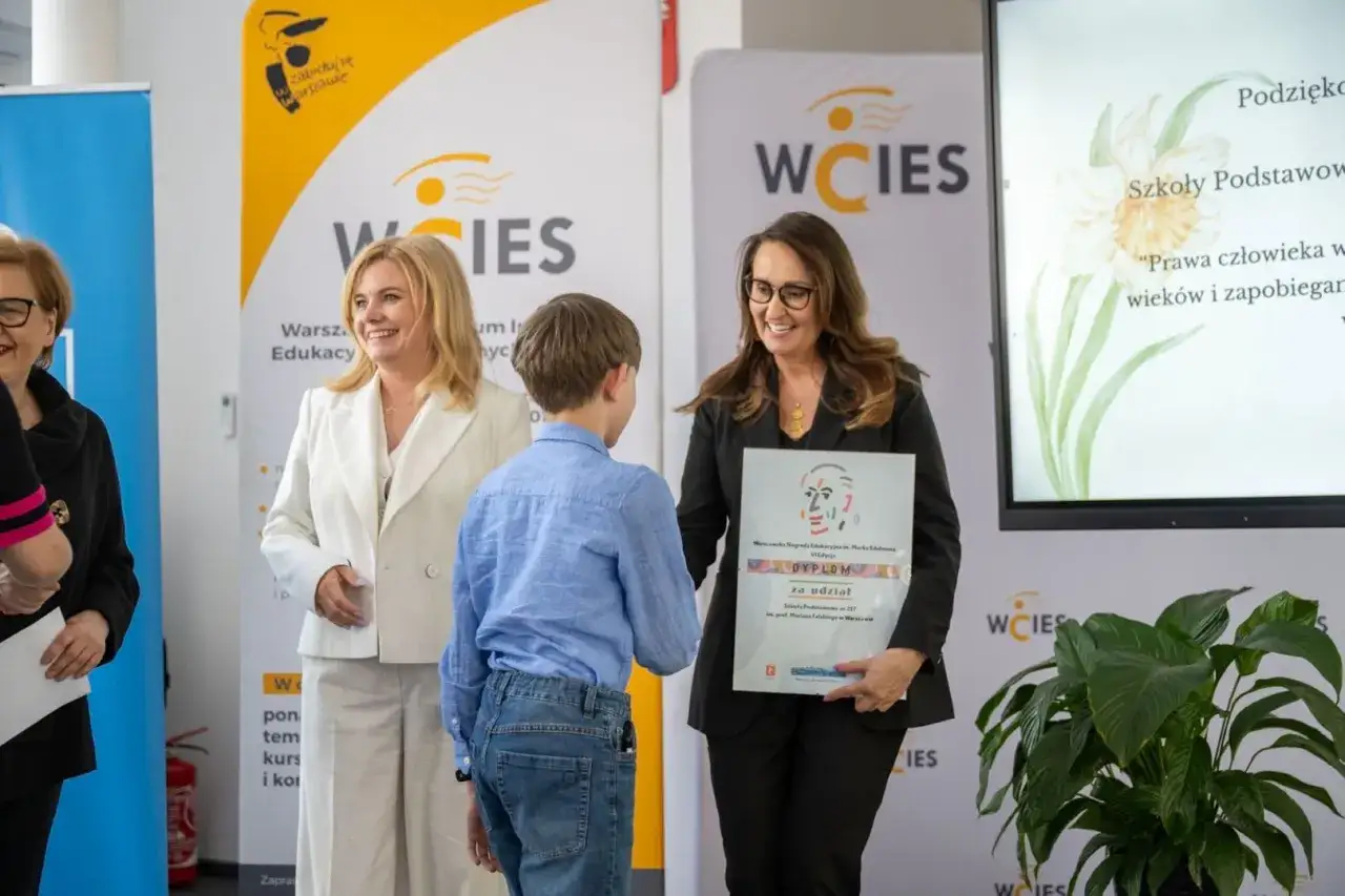 10 grudnia. Chłopiec odbiera dyplom od kobiety w czarnej marynarce. Obok stoi kobieta w białym żakiecie.