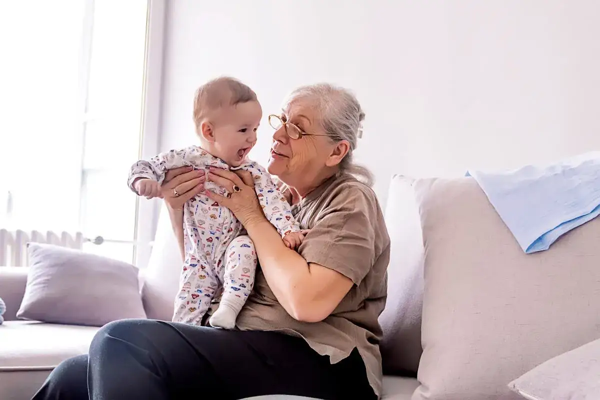 Babciowe ile wynosi? Uśmiechnięta babcia trzyma na rękach niemowlę w pajacyku z autkami.