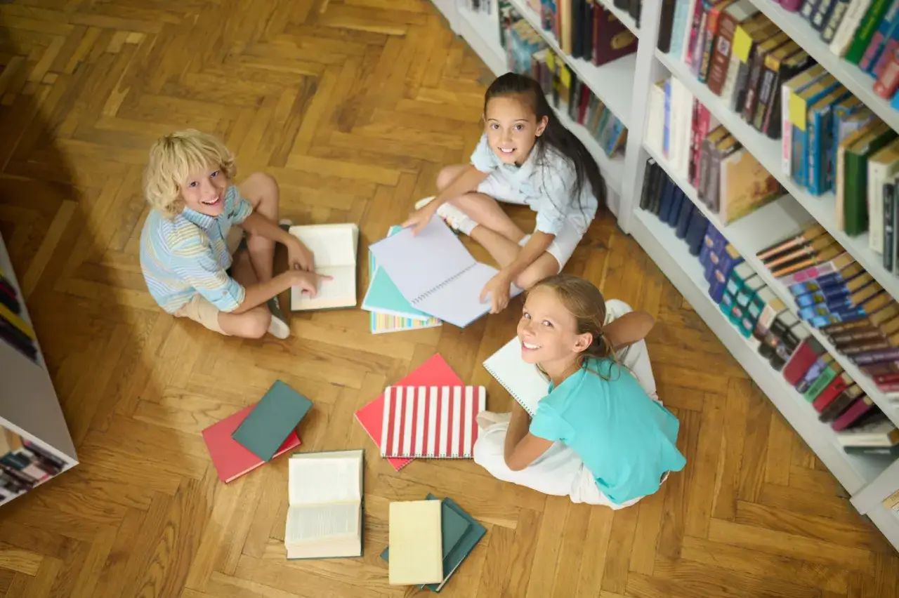 Dzieci uczą się czytać przy regałach z książkami. 23 kwietnia to dzień, który warto poświęcić na rozwijanie pasji do literatury.