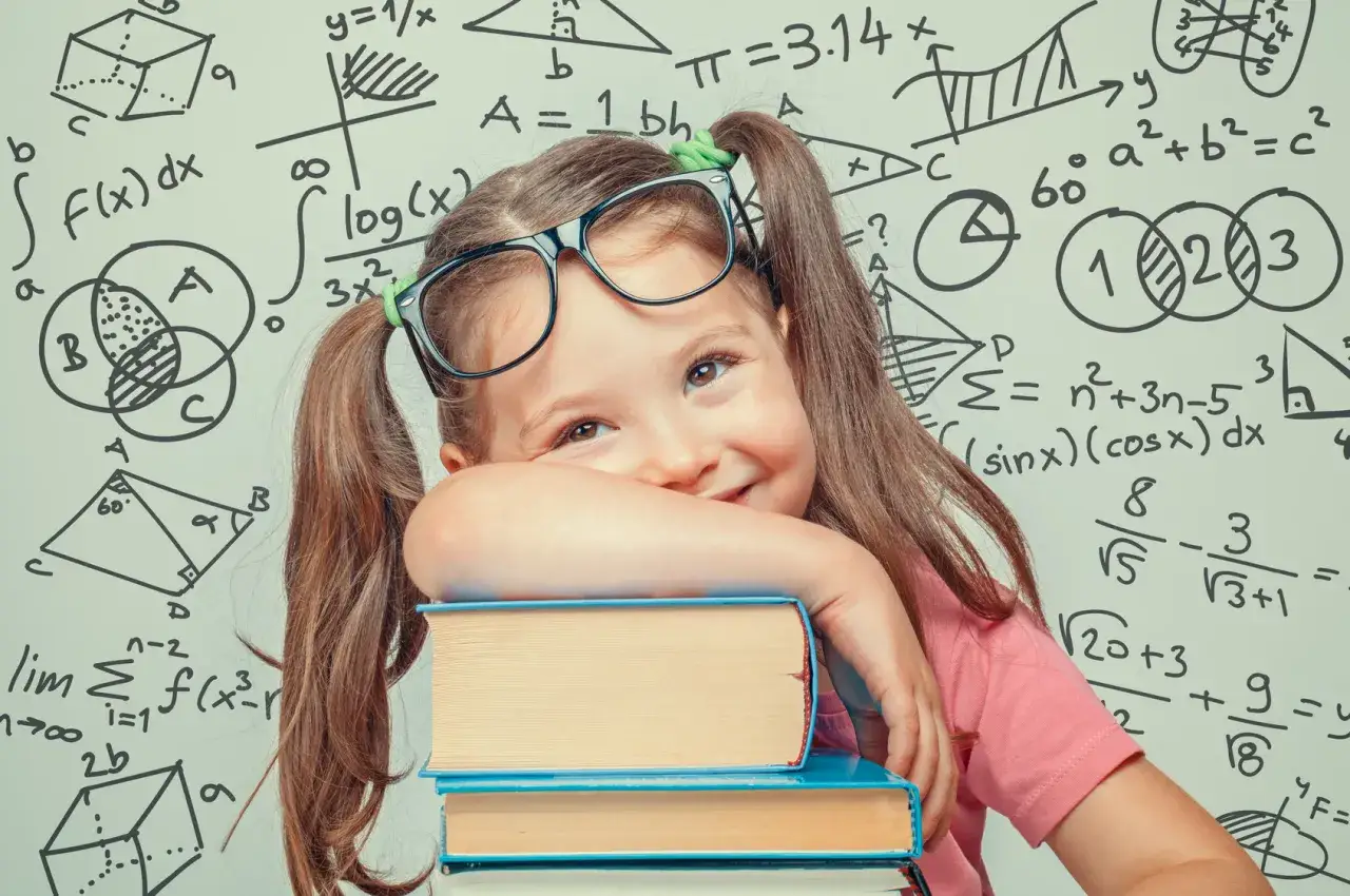Mała dziewczynka w okularach opiera się o stos książek na tle tablicy z matematycznymi wzorami. 12 marca nietypowe święta dla małego geniusza.