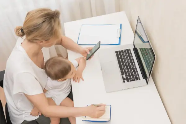 Kobieta z dzieckiem na kolanach sprawdza w telefonie, ile płatny urlop macierzyński. Obok laptop i notatnik.