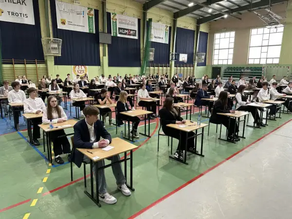 Uczniowie siedzą przy ławkach w sali gimnastycznej, skupieni na zadaniach. To ważny dzień, kiedy egzamin ósmoklasisty.