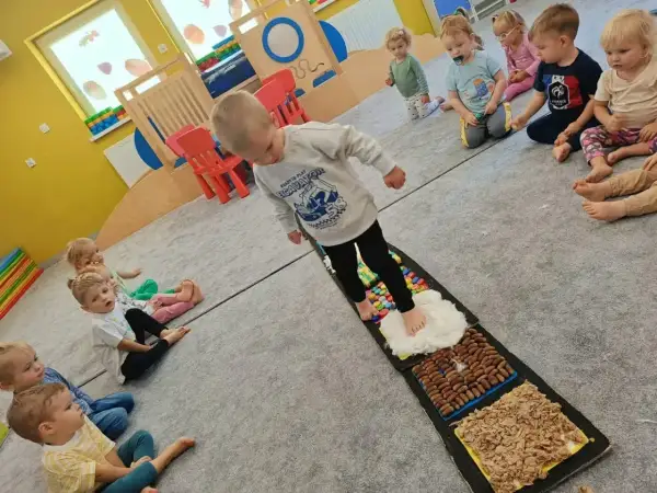 Dzieci aktywnie w żłobku ćwiczą zmysły na ścieżce sensorycznej. Maluchy z zaciekawieniem obserwują kolegę stąpającego po różnych fakturach.