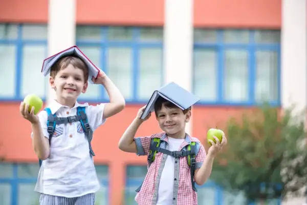 Dwóch chłopców z książkami na głowach i jabłkami w rękach przed szkołą. Zastanawiam się, czy to już 6 klasa, ile lat mają ci mali uczniowie?