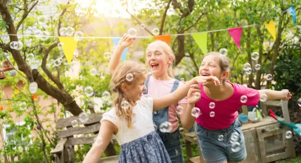 Trzy dziewczynki śmieją się i łapią bańki mydlane w ogrodzie. Wesołe życzenia urodzinowe dla dziecka!