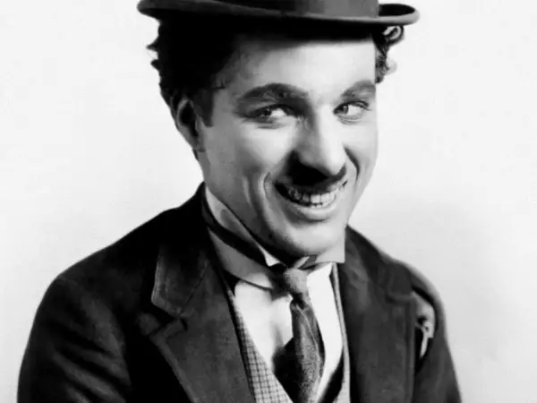 Charlie Chaplin w meloniku i garniturze, z uśmiechem, jakby opowiadał dowcip swoim dzieciom.