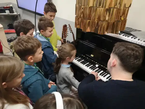 Dzieci słuchają lekcji muzyki przy pianinie. Nauczyciel pokazuje, do której klasy jest muzyka, a mali melomani z uwagą obserwują.