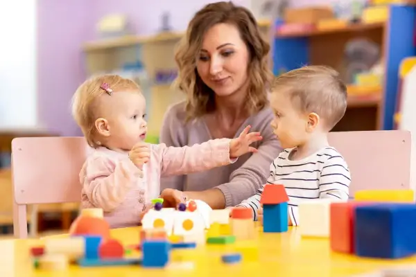 Dzieci bawią się klockami przy stole, a opiekunka patrzy na nich z uśmiechem. Dofinansowanie do żłobka ułatwia rodzicom opiekę nad maluchami.